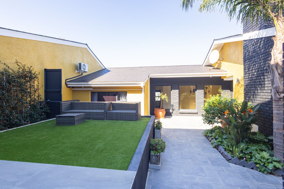 A well-maintained rear courtyard featuring a paved patio area, artificial lawn, and modern exterior finishes with contrasting yellow and dark brick walls.