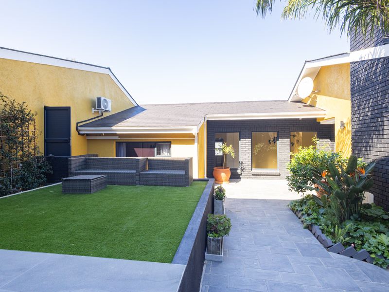 A well-maintained rear courtyard featuring a paved patio area, artificial lawn, and modern exterior finishes with contrasting yellow and dark brick walls.