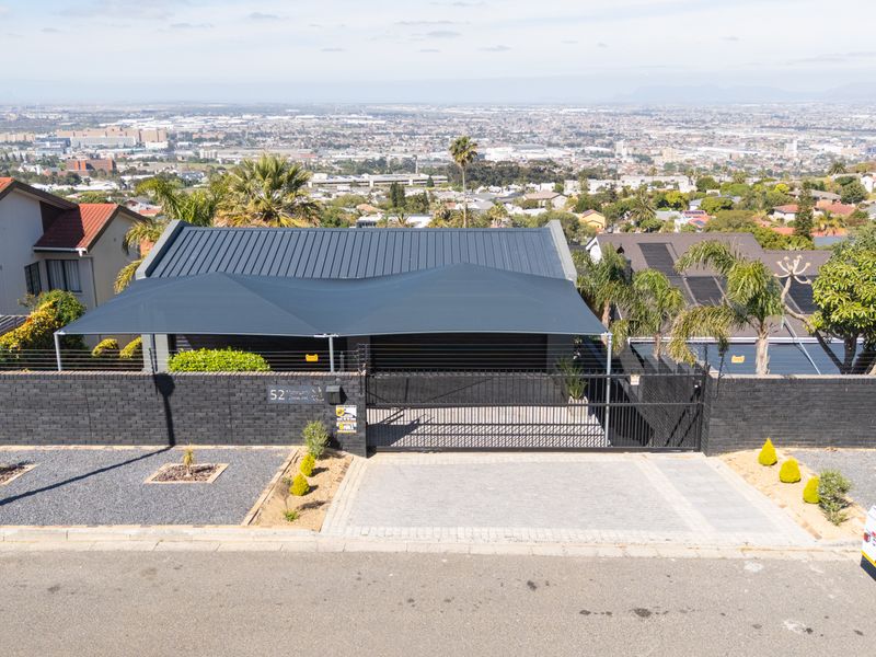 Aerial view of the property frontage featuring a paved driveway, secure gated entrance, brick boundary wall with electric fencing, and a modern shade sail.