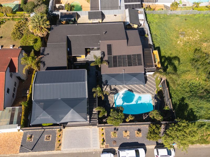 Aerial view of a residential property featuring a swimming pool, solar panels, paved driveway, and landscaped outdoor areas with shade structures.