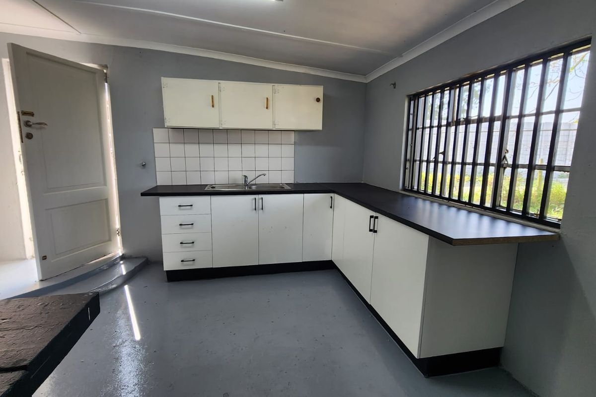 A functional, empty kitchen featuring L-shaped white cabinetry, a stainless steel sink, tiled backsplash, and a window with security bars.