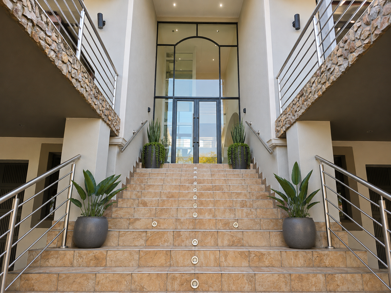 Grand entrance featuring a wide tiled staircase with stainless steel handrails, stone-clad pillars, and a modern glass door entry.