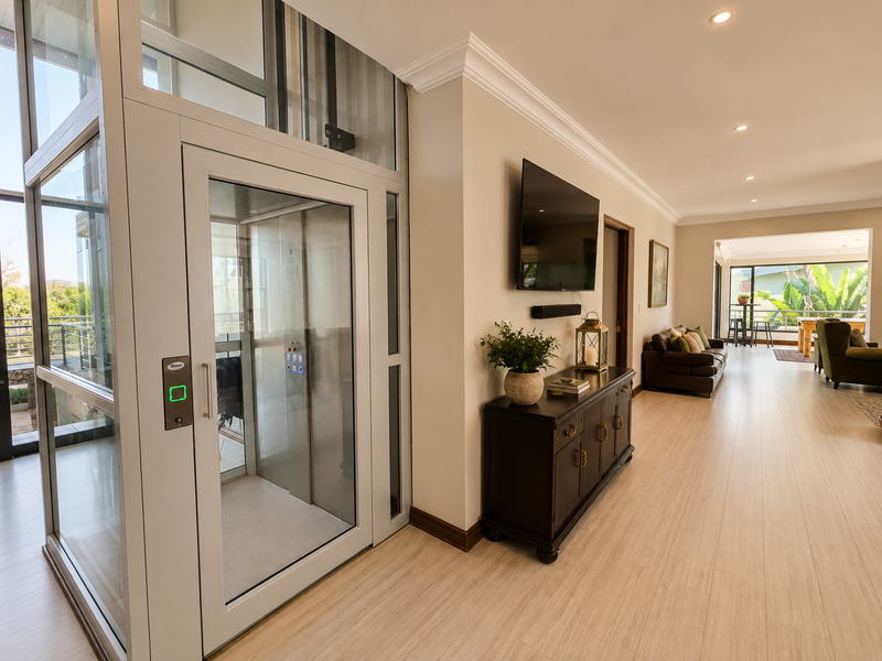 Spacious living area featuring a modern glass elevator, recessed lighting, crown molding, and light-toned laminate flooring, leading to an open-plan lounge space.
