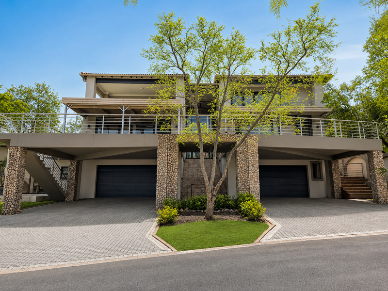 Modern double-story residence featuring a paved driveway, double garage, stone-clad pillars, and an expansive upper-level balcony with stainless steel railings.