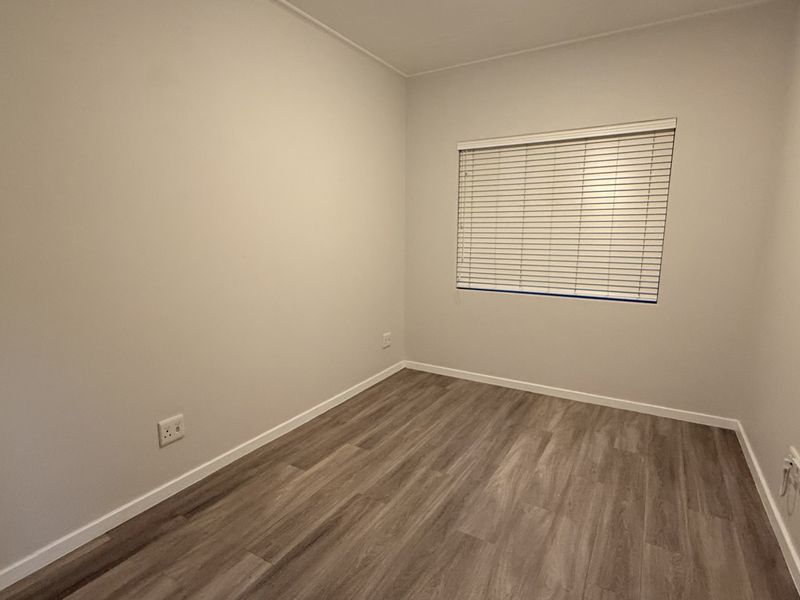 A clean, empty bedroom featuring modern laminate flooring, neutral wall paint, and fitted venetian blinds.