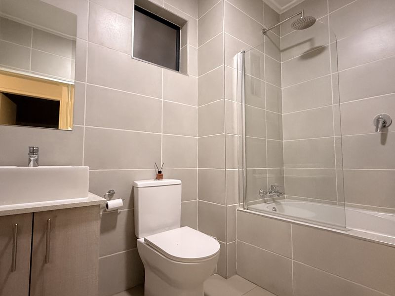 Modern bathroom featuring neutral wall tiles, a vessel sink vanity, a toilet, and a bathtub with a glass shower screen.