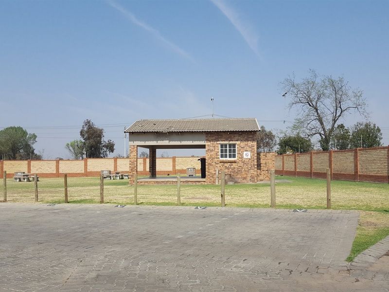 A communal outdoor pavilion with a brick structure and tiled roof, situated within a fenced complex with paved parking and lawn area.