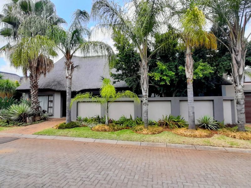 Front elevation of a property featuring a traditional thatched roof, a secure boundary wall, a roller garage door, and a well-maintained landscaped garden.