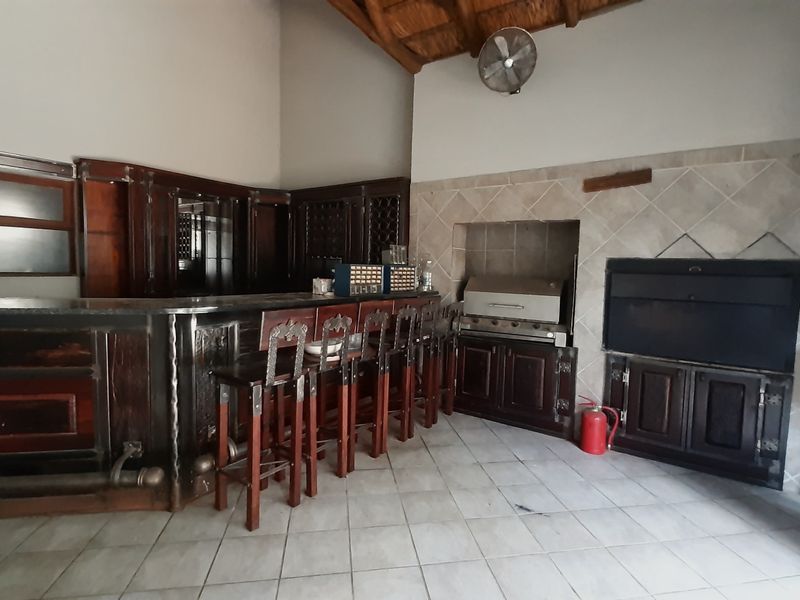 A spacious indoor entertainment area featuring a large built-in wooden bar, integrated braai, tiled flooring, and high ceilings with exposed timber rafters.