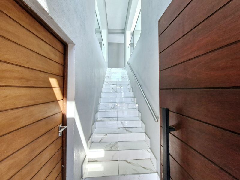A modern, bright staircase featuring marble-look tiled steps, a sleek metal handrail, and a glass balustrade, flanked by timber doors.