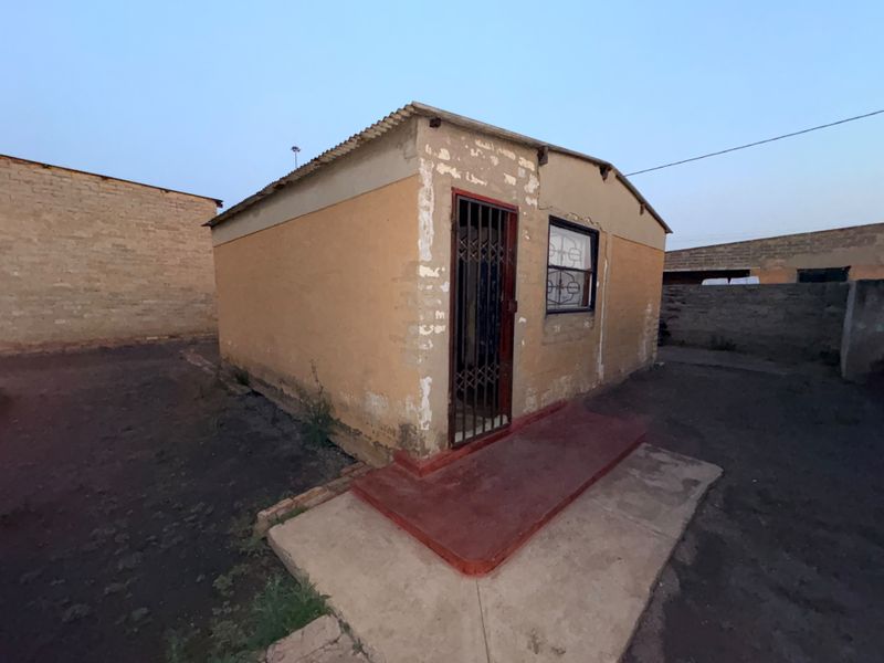 Exterior view of a small residential structure featuring a corrugated iron roof, security gate, and window bars, set on a compact plot.