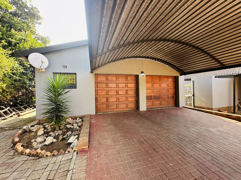 A double garage featuring wooden doors, protected by a large curved corrugated metal carport, with a paved driveway and landscaped garden bed.