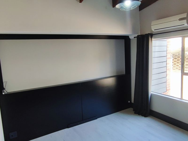 A bright, empty bedroom featuring modern black wall paneling, exposed timber ceiling beams, laminate flooring, and a wall-mounted split-system air conditioner.
