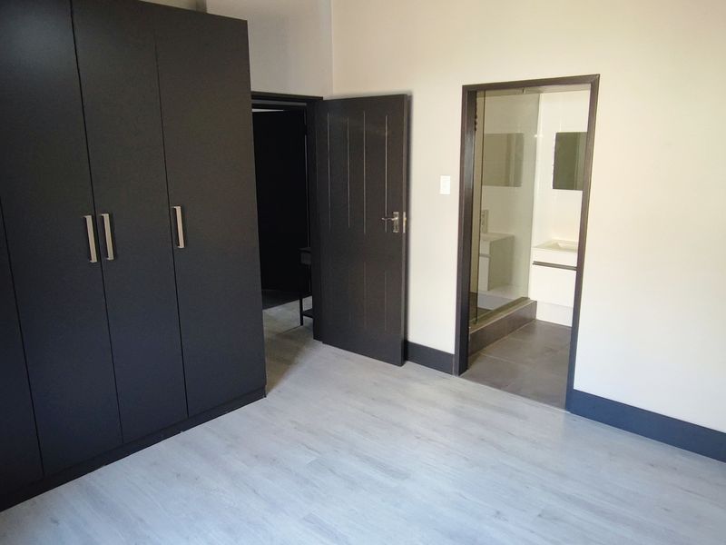 A clean, modern bedroom featuring light laminate flooring, dark built-in cupboards, and direct access to an en-suite bathroom.