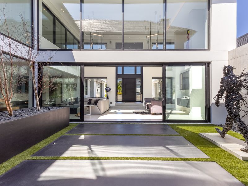 Modern rear exterior featuring a double-volume glass facade, large sliding doors, concrete paving, and a landscaped courtyard area.