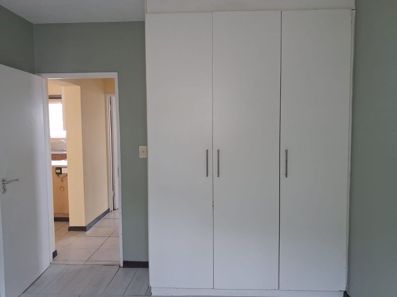 A bedroom featuring a large white built-in wardrobe, neutral wall paint, and tiled flooring, with a view into the adjacent kitchen area.