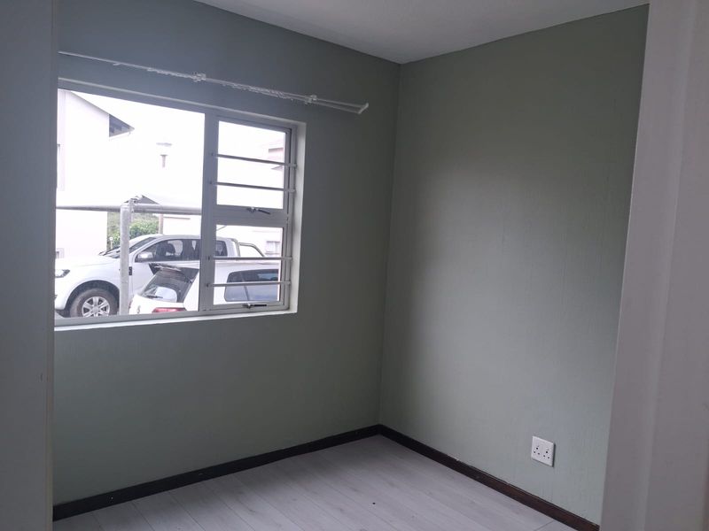 An empty bedroom featuring light-toned laminate flooring, sage green walls, and an aluminium-framed window with security bars.