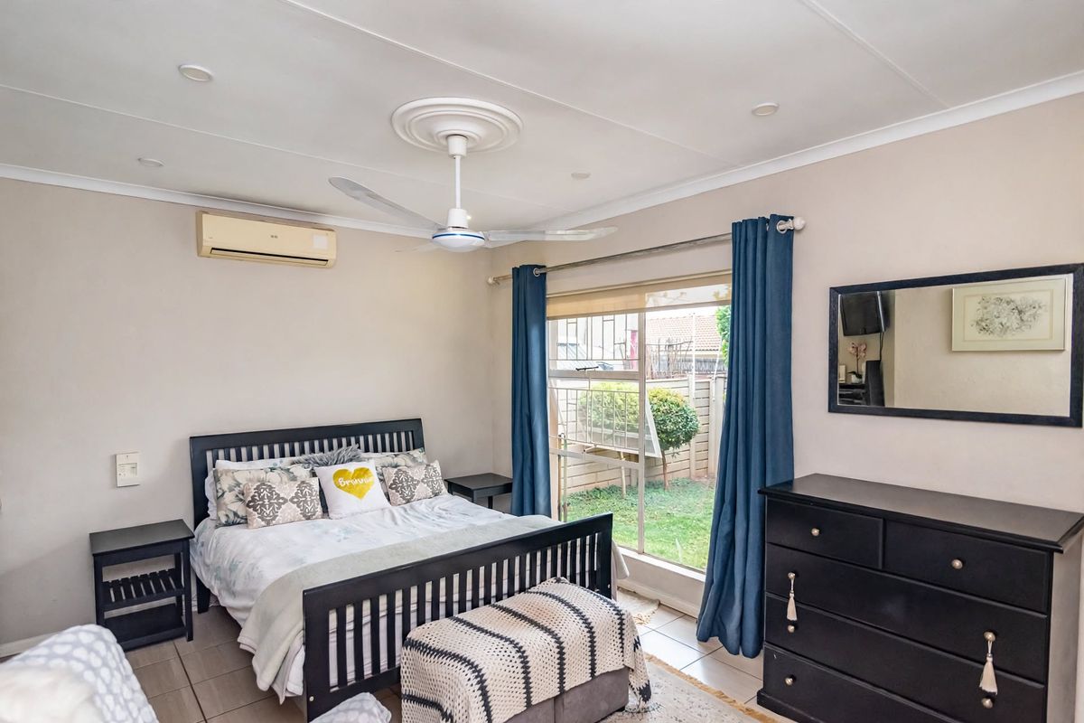 A well-lit bedroom featuring tiled flooring, a ceiling fan, and a wall-mounted air conditioning unit, with direct access to a small garden area.