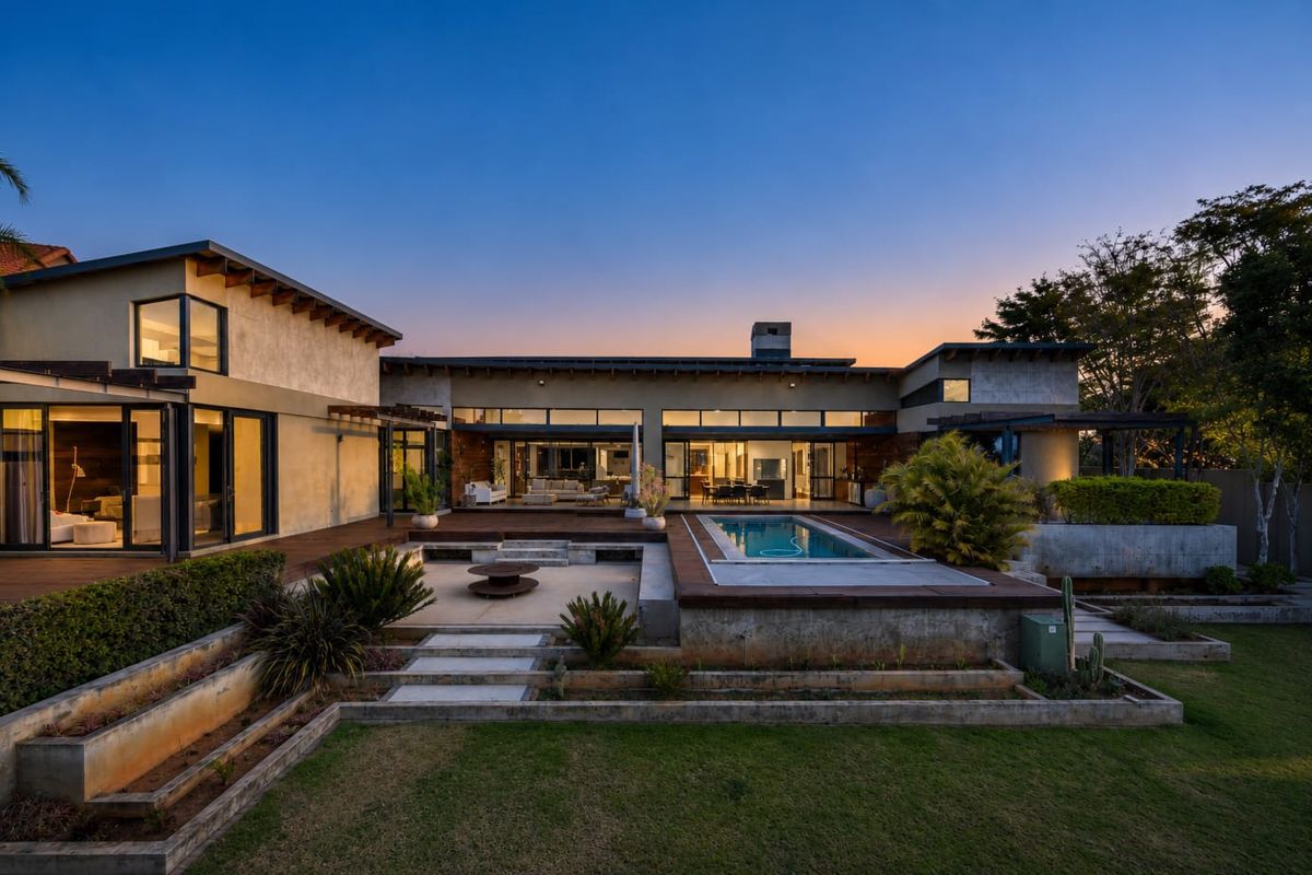 Stunning rear exterior view featuring a modern residence with timber decking, a swimming pool, and tiered landscaped gardens at dusk.
