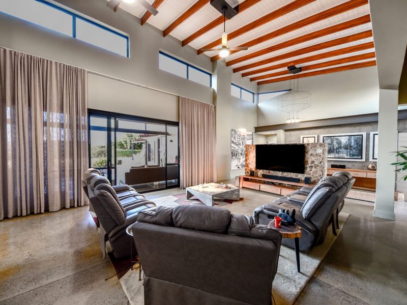 Spacious living room featuring high ceilings with exposed timber beams, polished concrete floors, a stone-clad feature wall, and large sliding doors leading to the patio.