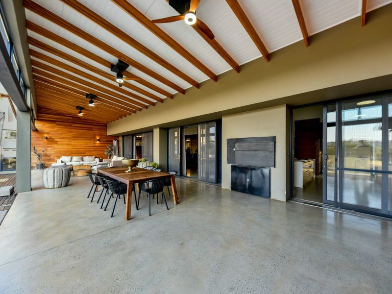 Spacious covered patio featuring a polished concrete floor, exposed timber beam ceiling with fans, timber wall cladding, and a built-in braai area.
