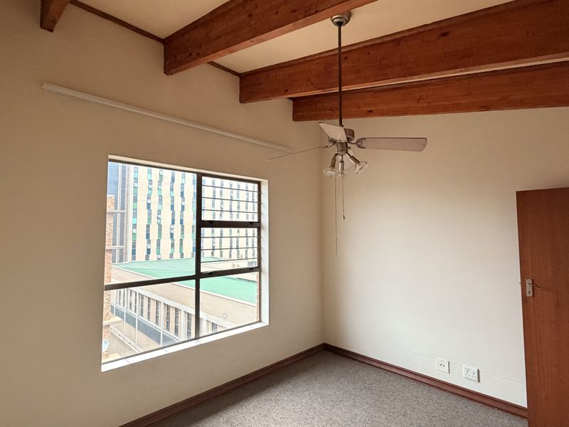 A bright, empty bedroom featuring exposed timber ceiling beams, a ceiling fan, carpeted flooring, and a large window with urban views.