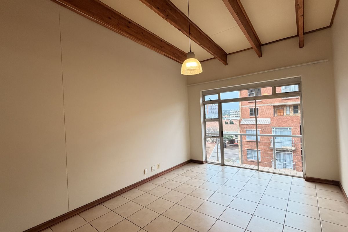 An empty living room featuring exposed timber ceiling beams, tiled flooring, and a large sliding glass door leading to a balcony.