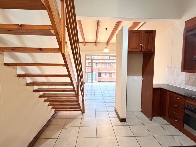 Open-plan living area featuring a wooden staircase, tiled flooring, exposed ceiling beams, and a functional kitchen with built-in cabinetry and appliances.