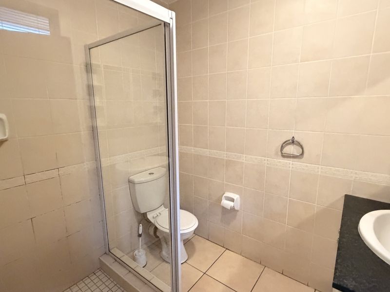 A functional bathroom featuring a glass shower enclosure, toilet, and vanity basin with neutral wall and floor tiling.