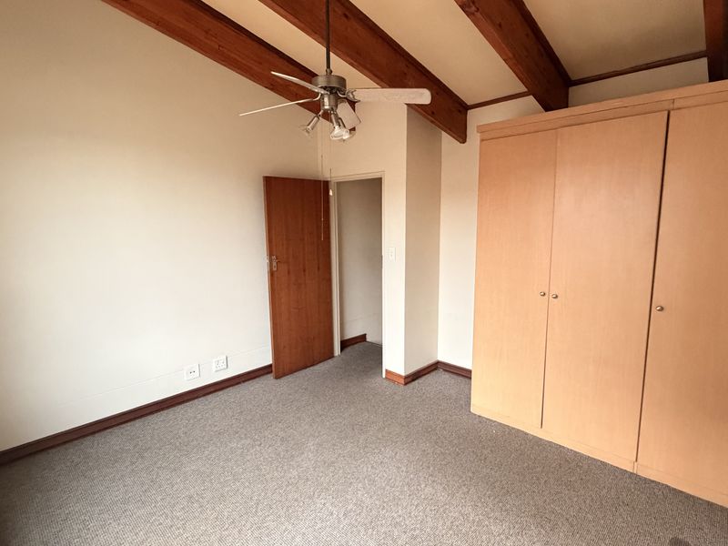 A spacious bedroom featuring exposed timber ceiling beams, a ceiling fan, neutral carpeting, and a large built-in wardrobe for storage.