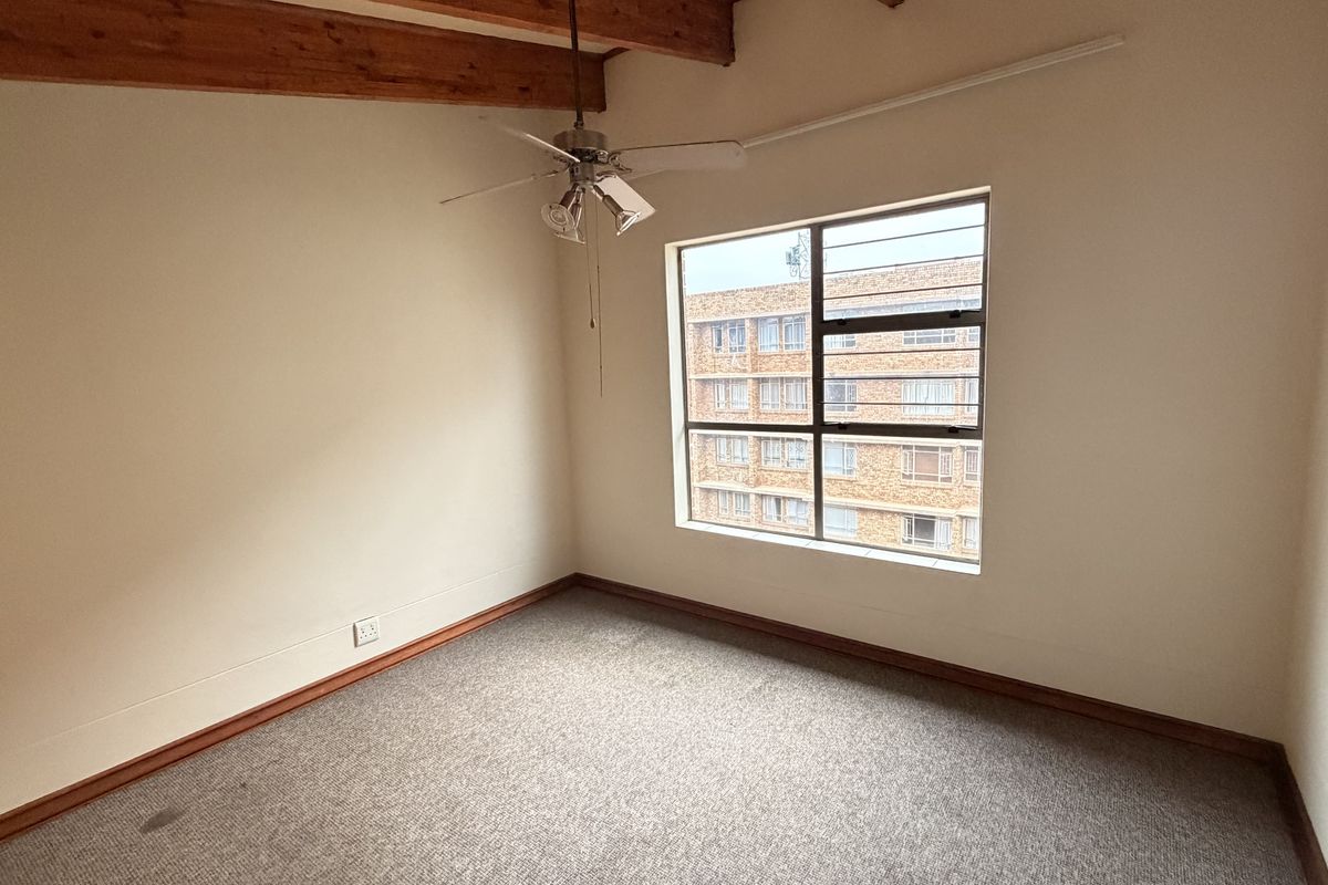 An empty bedroom featuring exposed timber ceiling beams, a ceiling fan, and a window with security bars.
