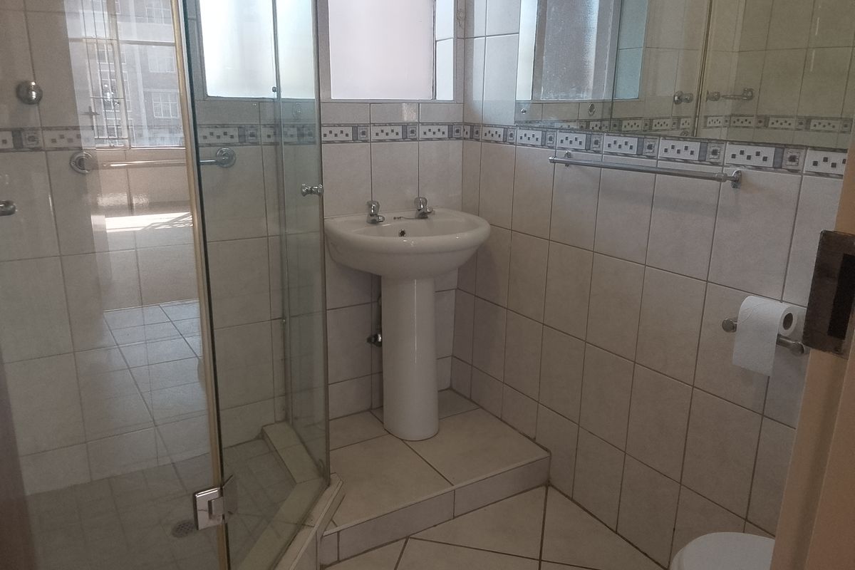 A functional bathroom featuring a pedestal basin, a glass-enclosed shower, and neutral wall and floor tiling.