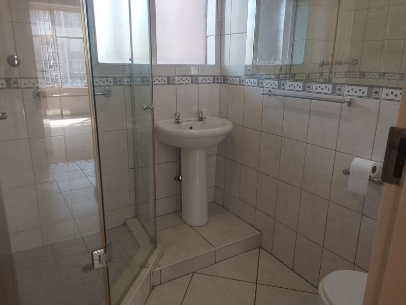 A functional bathroom featuring a pedestal basin, a glass-enclosed shower, and neutral wall and floor tiling.
