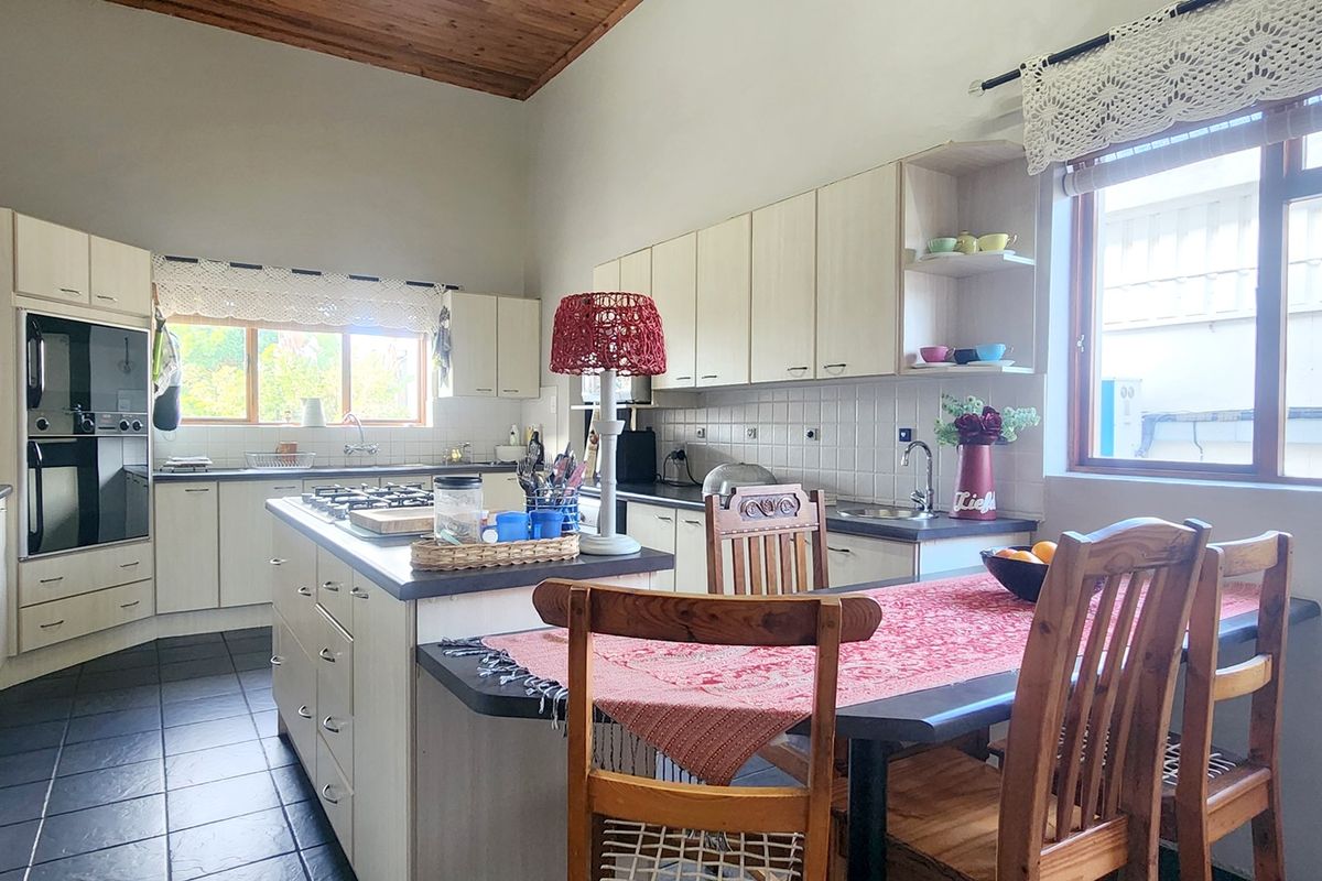 A functional kitchen featuring light-toned cabinetry, a central island with a gas hob, built-in oven, and dark tiled flooring under a timber-paneled ceiling.