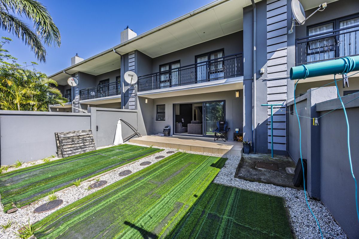 Rear view of the townhouse featuring a tiled patio, small garden area with artificial turf, and an upper-level balcony with metal railings.