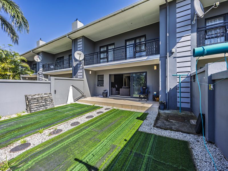 Rear view of the townhouse featuring a tiled patio, small garden area with artificial turf, and an upper-level balcony with metal railings.