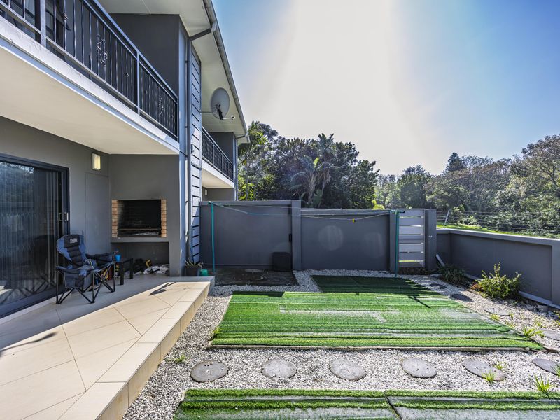 Rear exterior view featuring a tiled patio with a built-in braai, balcony access, and a low-maintenance garden area with boundary walling.