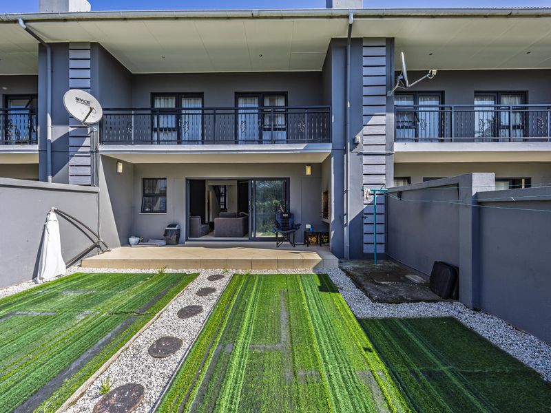 Rear view of a townhouse featuring a tiled patio, small garden with artificial lawn, and an upper-level balcony with metal railings.