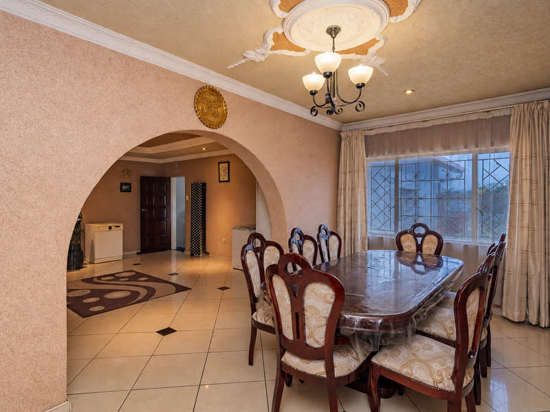 A formal dining room featuring an arched entryway, decorative ceiling rose with chandelier, and tiled flooring, connecting to an adjacent living space.