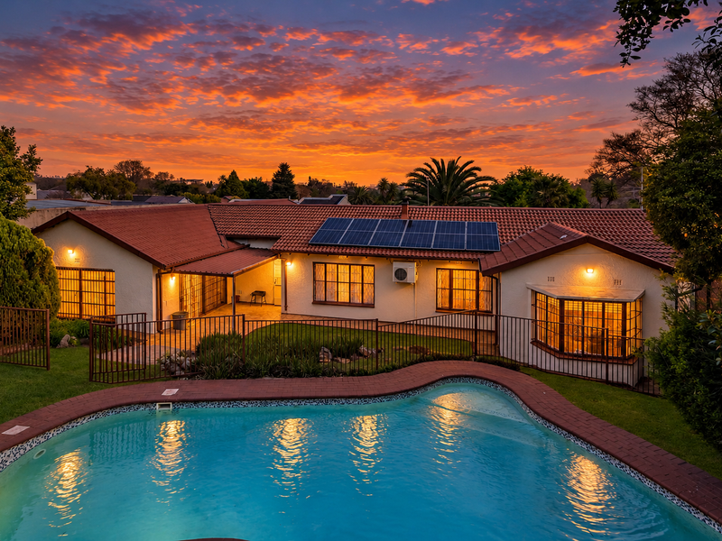 Rear view of the house featuring a swimming pool, landscaped garden, solar panels, and a covered patio area at sunset.