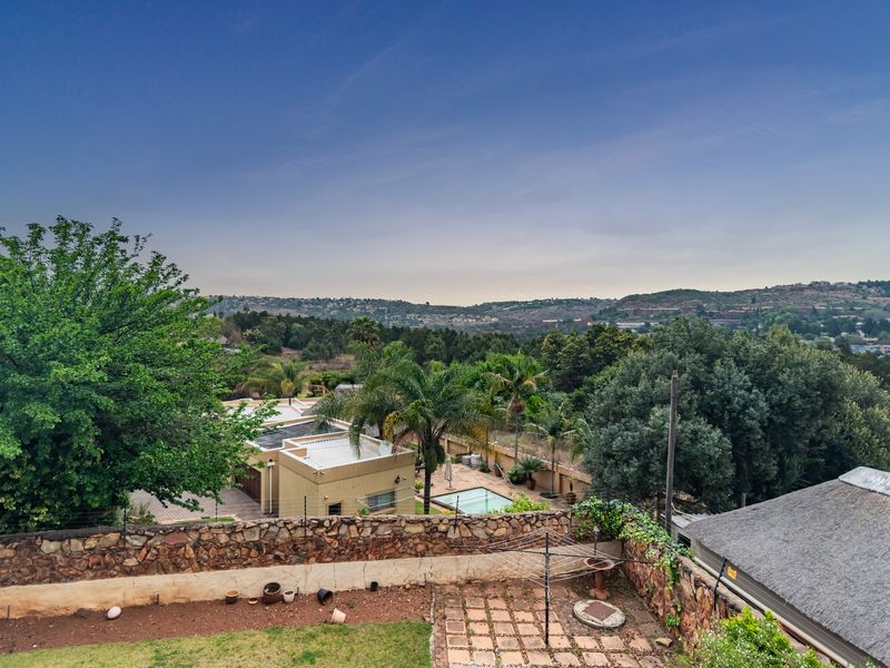 Elevated view overlooking a residential property featuring a swimming pool, stone boundary wall, and surrounding landscape with distant hills.