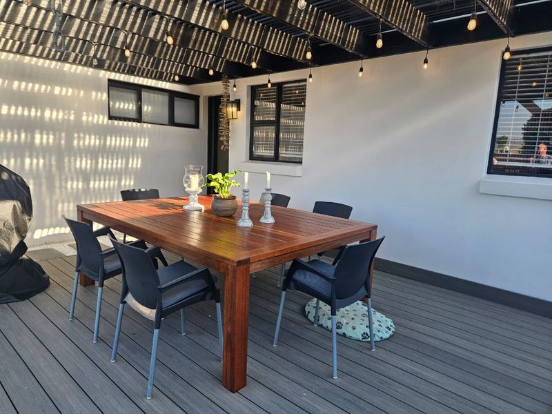 A well-maintained outdoor patio area featuring timber decking, a covered pergola with string lighting, and modern aluminium-framed windows.