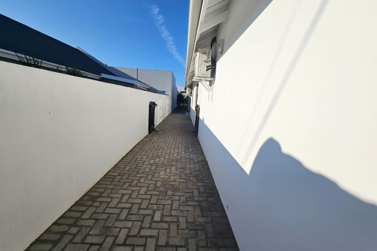 A narrow paved side passage or walkway between two white boundary walls, featuring an external air conditioning unit mounted on the building exterior.
