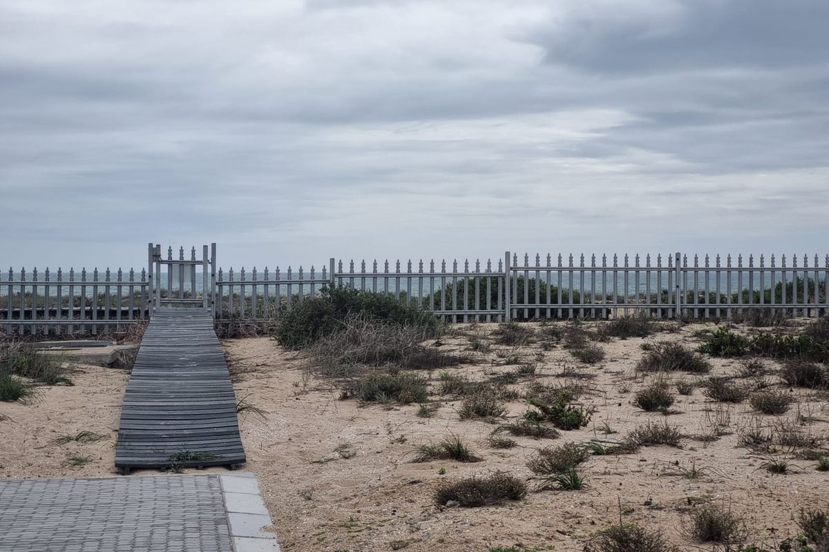 A rear exterior view of the property featuring a sandy landscape, a wooden boardwalk leading to a metal gate, and a perimeter fence.