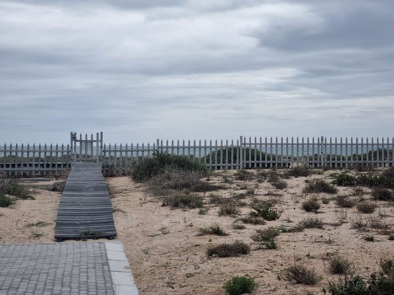 A rear exterior view of the property featuring a sandy landscape, a wooden boardwalk leading to a metal gate, and a perimeter fence.