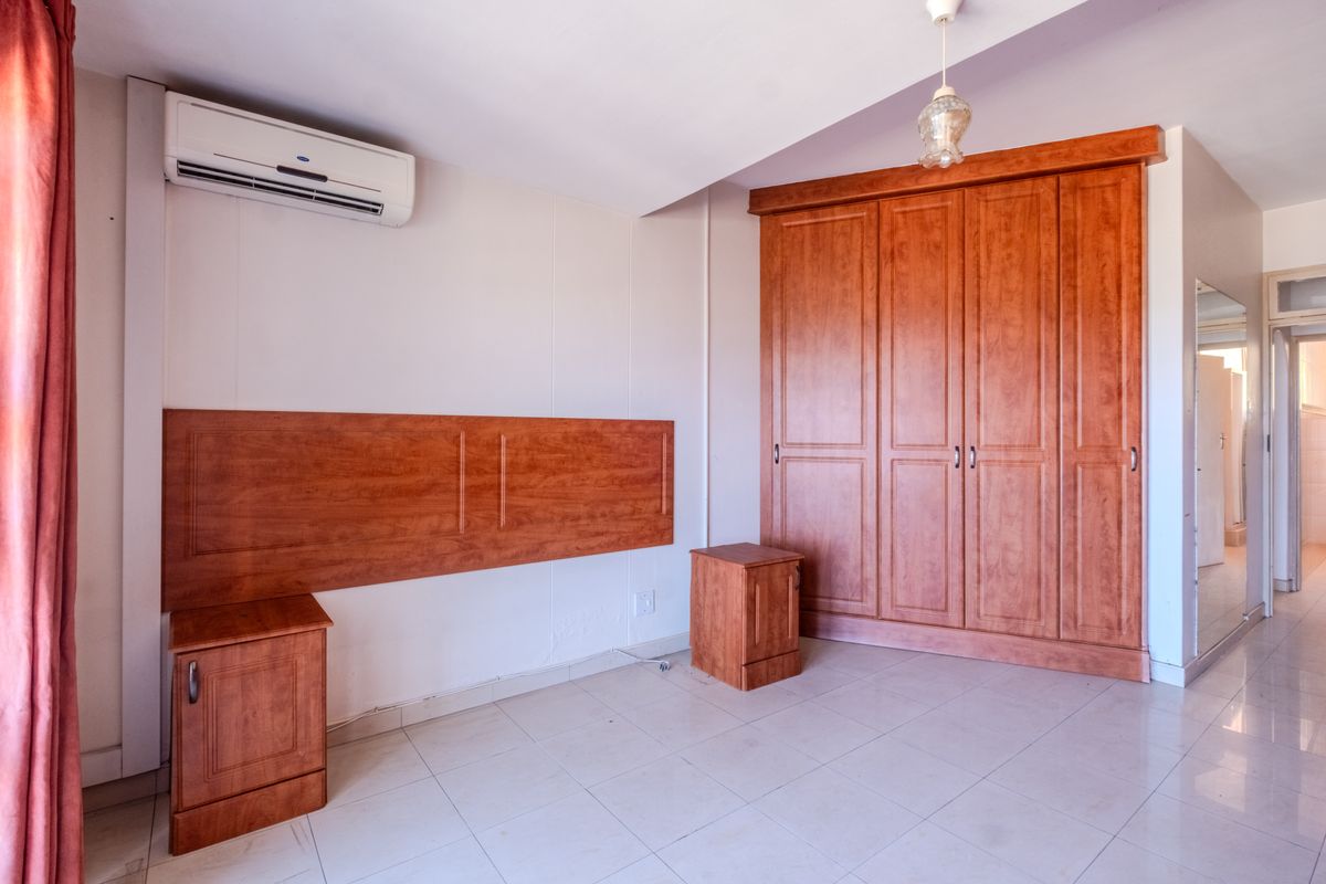 A spacious bedroom featuring tiled flooring, a large built-in wooden wardrobe, a matching built-in headboard, and a wall-mounted air conditioning unit.