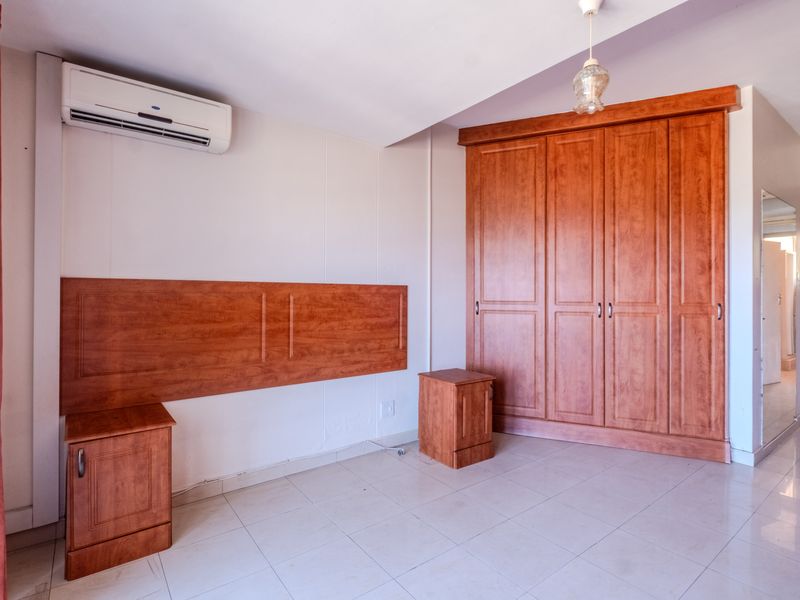A spacious bedroom featuring tiled flooring, a large built-in wooden wardrobe, a matching built-in headboard, and a wall-mounted air conditioning unit.