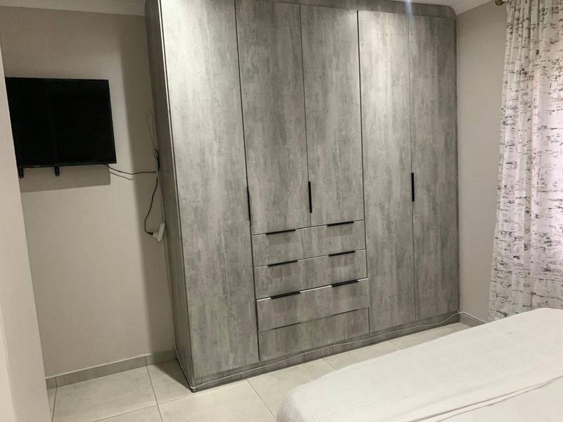 A bedroom featuring modern grey wood-grain built-in cupboards with integrated drawers and neutral floor tiles.