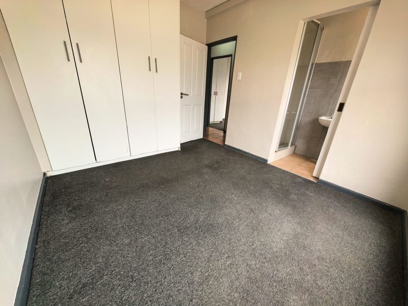 An empty bedroom featuring built-in cupboards, carpeted flooring, and an attached en-suite bathroom with a shower and pedestal basin.