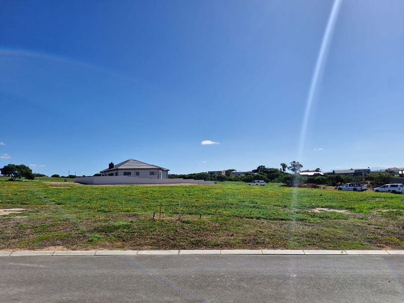 A vacant residential plot of land featuring a level grassy surface, bordered by a paved road and kerb in a developing neighbourhood.